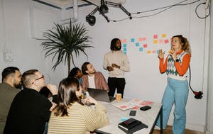 Team brainstorming around a table while a colleague presents beside a wall of colorful sticky notes.