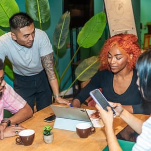 A group of four coworkers gathers around a table, reviewing information on a tablet and smartphone in a modern office.