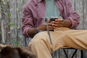 A person sits outdoors in orange-striped pants and a light magenta jacket while holding a smartphone.