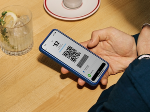 A hand holds a blue smartphone displaying the PayPal app's Pay screen with a ¥52 QR code and barcode, ready to show for payment at a restaurant table with a garnished cocktail and place setting in the background.