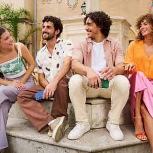 Four friends sit outdoors near a fountain, smiling and talking together on a sunny day.