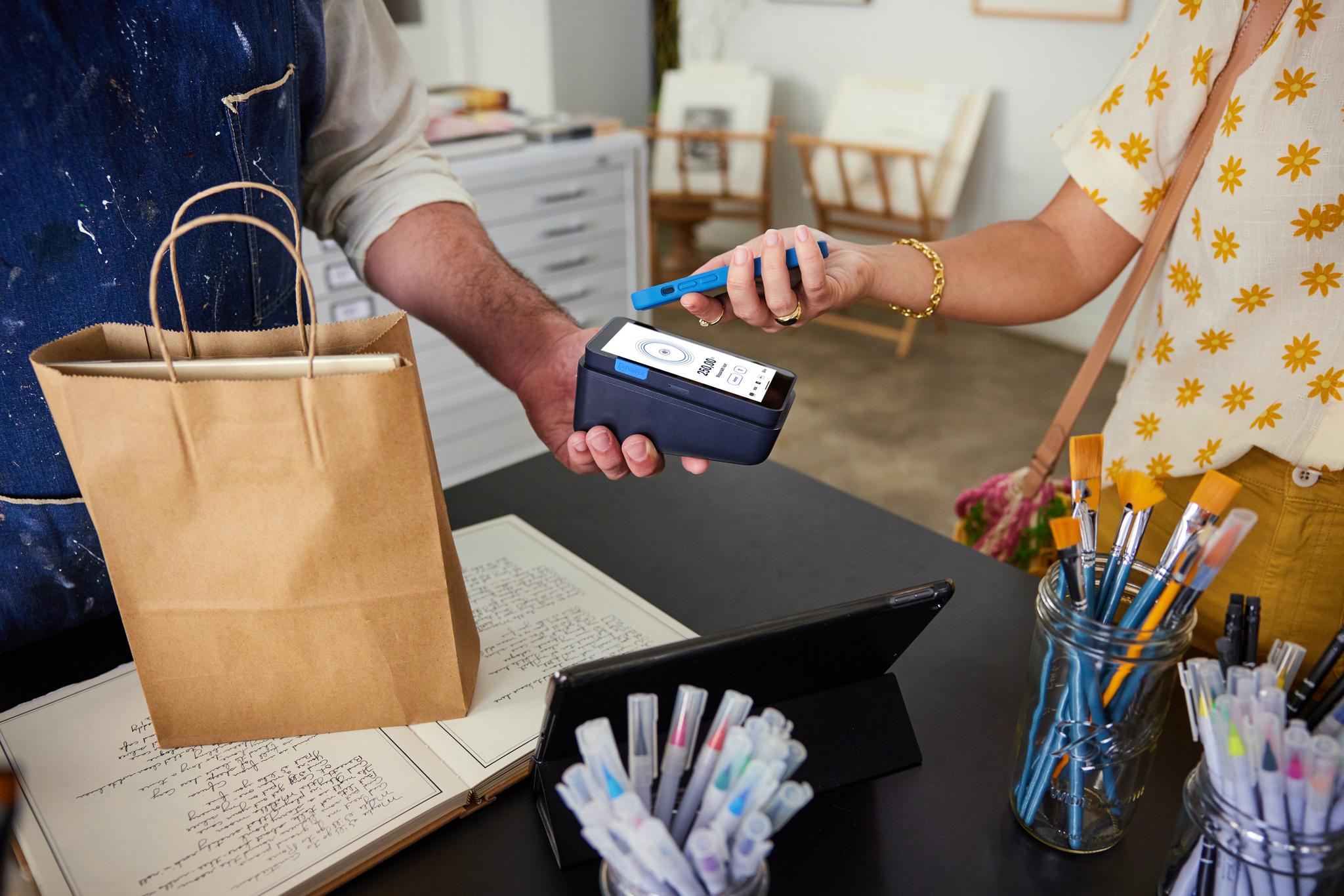 A shop employee holding a PayPal Terminal in one hand accepting a contactless payment from customer’s phone